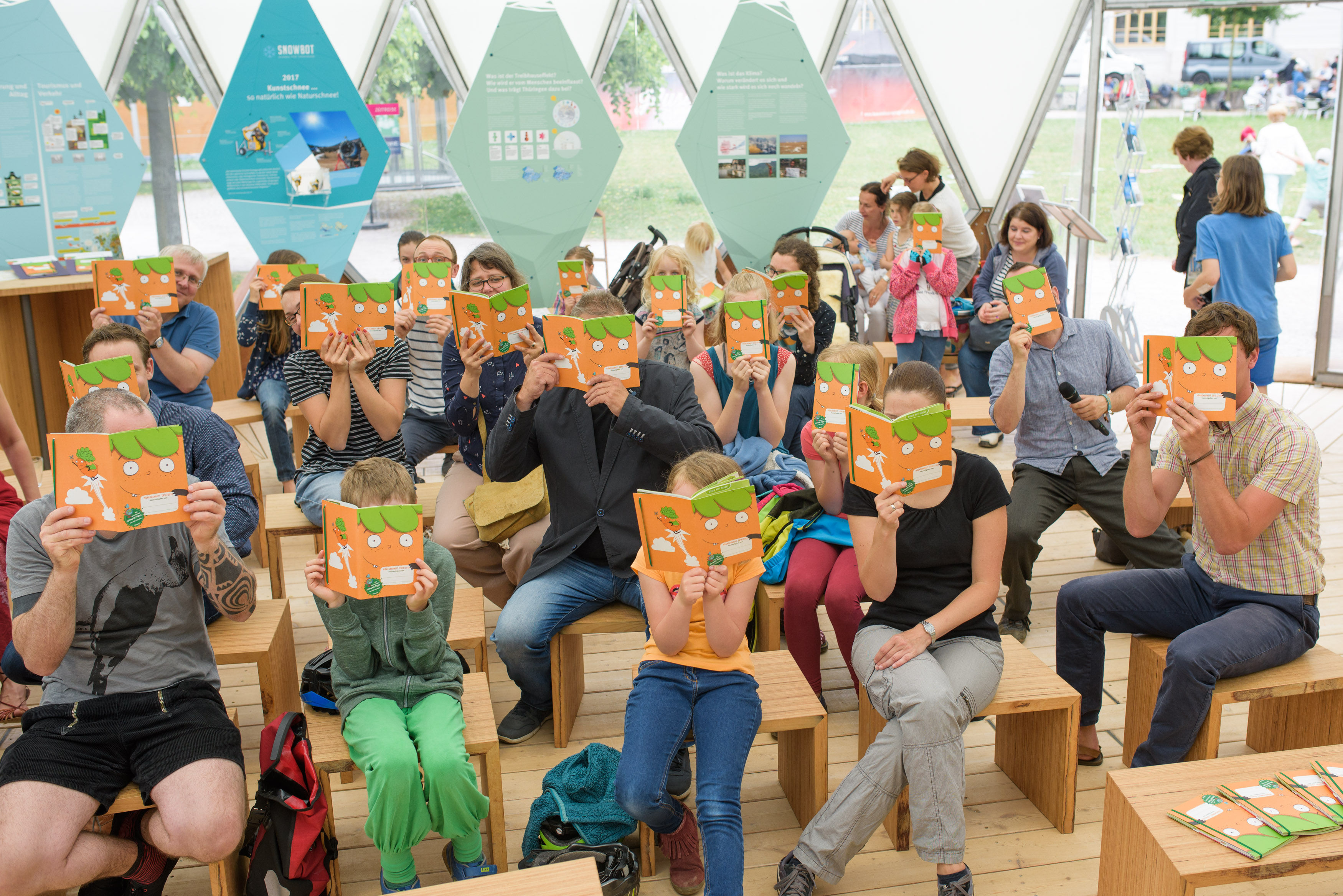 Erwachsene und Kinder sitzen im Klimapavillon und halten aufgeschlagene Möhrchenhefte vor ihre Gesichter